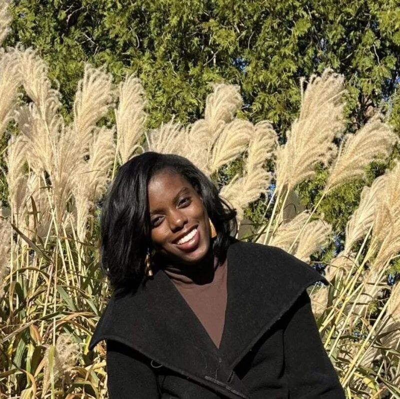 The image shows a smiling Black woman in front of tall, feathery grasses. She is wearing a black coat over a brown top. The background features green foliage, suggesting an outdoor setting, possibly during autumn. The woman's expression is warm and inviting.